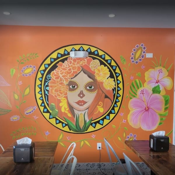 Vibrant Mexican-themed mural with a Day of the Dead woman, flowers, and corn on an orange wall.