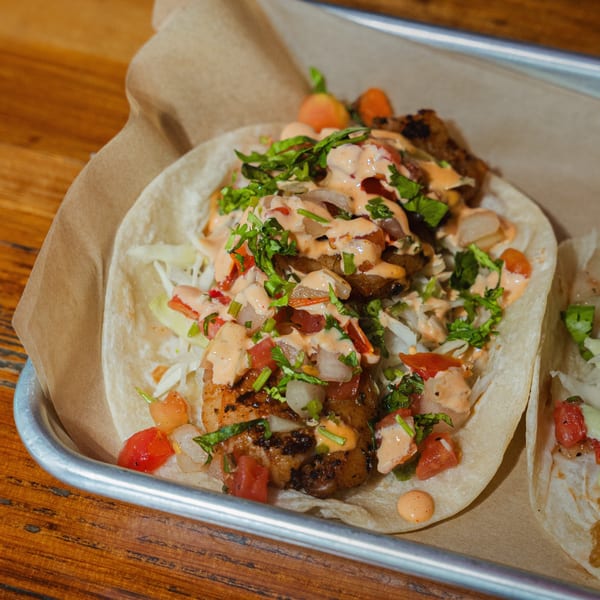 Two delicious Taco Station tacos with grilled shrimp, salsa, and creamy sauce on a metal tray.