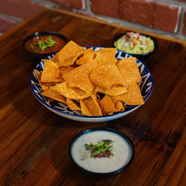 A bowl of tortilla chips with salsa, guacamole, and queso dip on a wooden table.