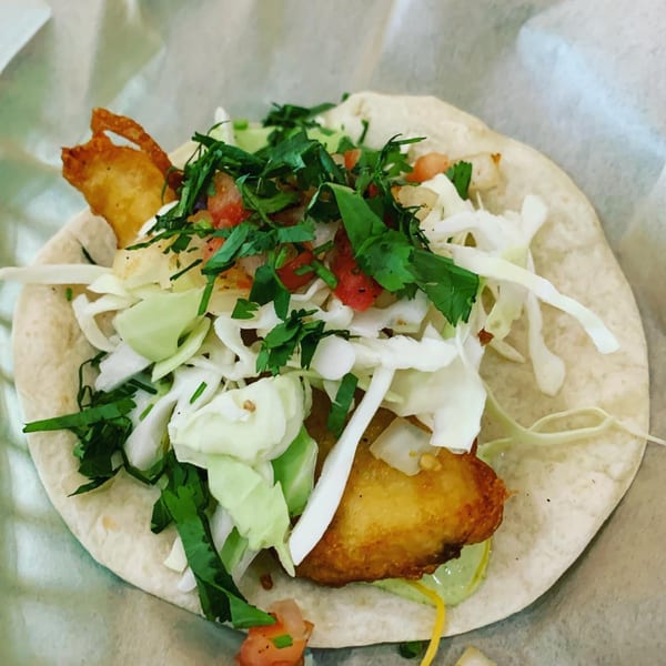 A close-up of a Taco Station fish taco with fried fish, cabbage, salsa, and cilantro.