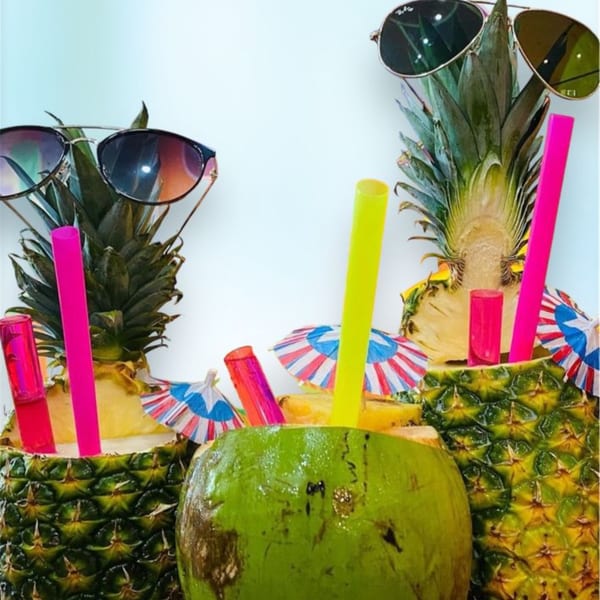 Two pineapples with sunglasses and a coconut drink with colorful straws and umbrellas.