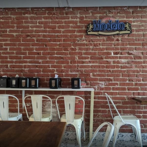 A Modelo sign hangs above a shelf with napkin dispensers against a brick wall at Taco Station.