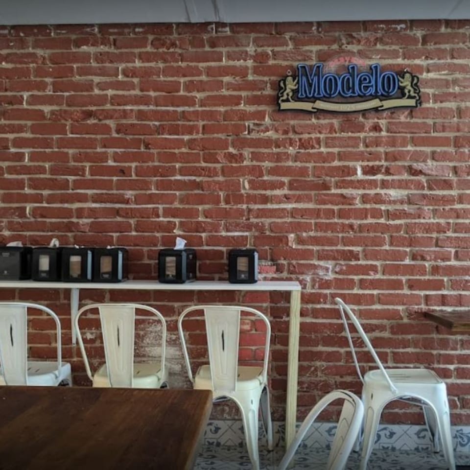 A Modelo sign hangs above a shelf with napkin dispensers against a brick wall at Taco Station.